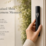 Hand holding a Timeless Raised Silver Shin Black Cement Mezuzah case with a smooth matte black surface and prominent sculptural silver-tone Hebrew Shin, showing the strong black and silver contrast in a contemporary neutral home setting