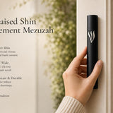 Hand holding a Timeless Raised Silver Shin Black Cement Mezuzah case with a smooth matte black surface and prominent sculptural silver-tone Hebrew Shin, showing the strong black and silver contrast in a contemporary neutral home setting