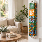 A colorful decorative mezuzah case mounted on a white door in a bright, modern living room. The case features a vibrant mosaic tile pattern in blue, green, orange, and yellow, with a prominent orange panel displaying the Hebrew letter Shin in white