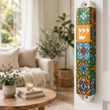 A colorful decorative mezuzah case mounted on a white door in a bright, modern living room. The case features a vibrant mosaic tile pattern in blue, green, orange, and yellow, with a prominent orange panel displaying the Hebrew letter Shin in white