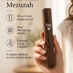 Hand holding a traditional woodgrain acrylic mezuzah case featuring a silver Hebrew Shin in a warm, minimalist space.