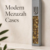 Close product view of a unique gray cement mezuzah case with a raised silver-tone Shin and long decorative inlay filled with golden amber, honey, bronze, and warm crystal and glass tones, styled against a clean contemporary background on a stone surface