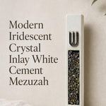 Close portrait of a unique modern white cement mezuzah case featuring a raised silver-tone Shin above a vertical panel of iridescent crystal and glass pieces with pearl, charcoal, bronze, green, and blue-gray reflections against a clean neutral backdrop