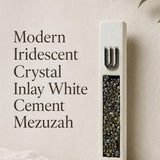 Close portrait of a unique modern white cement mezuzah case featuring a raised silver-tone Shin above a vertical panel of iridescent crystal and glass pieces with pearl, charcoal, bronze, green, and blue-gray reflections against a clean neutral backdrop