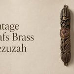 Vintage Leafs brass mezuzah case mounted on a neutral wall, featuring an aged antique brass finish, deeply sculpted leaves and botanical details, and a raised Hebrew Shin medallion at the center. This traditional brass mezuzah case brings warm vintage Judaica character to a Jewish home, entryway, or interior doorway