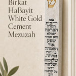 White cement Birkat HaBayit mezuzah displayed upright against a minimalist background, featuring a raised gold Shin and the full Hebrew home blessing in contrasting lettering across the front of the slim contemporary case
