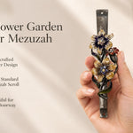 Wildflower Garden Pewter Mezuzah mezuzah cover held upright in one hand, showing yellow and purple flowers with sparkling centers, a small reddish blossom and green curling leaves along a textured silver-toned pewter stem beside the product name and illustrated design features in a wide social-sharing graphic for decorative Jewish home Judaica